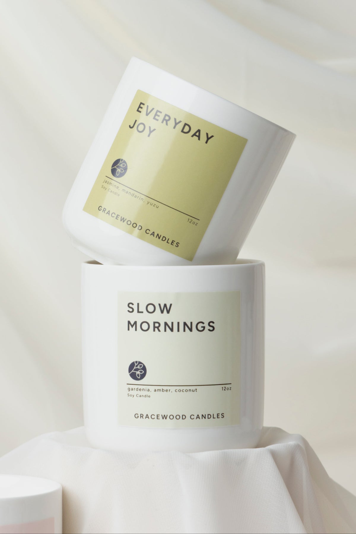 Two candles labeled 'Everyday Joy' and 'Slow Mornings' on a white fabric background with green leaves.