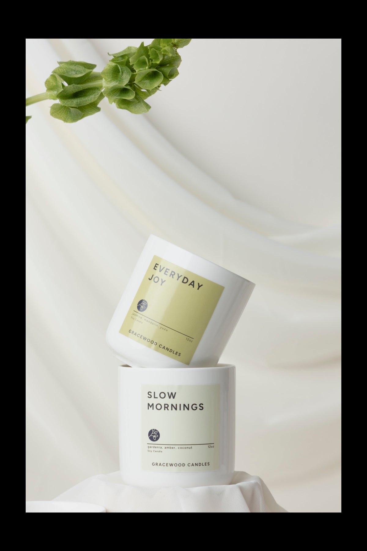 Two candles labeled 'Everyday Joy' and 'Slow Mornings' on a white fabric background with green leaves.