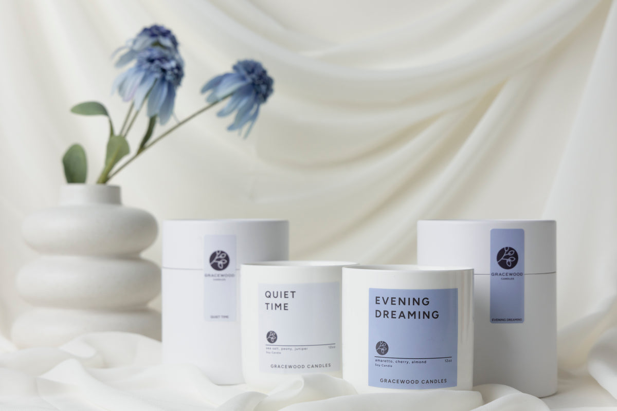 Three candles labeled 'Quiet Time', 'Evening Dreaming', and 'Wisteria' on a white fabric background with a vase of blue flowers.