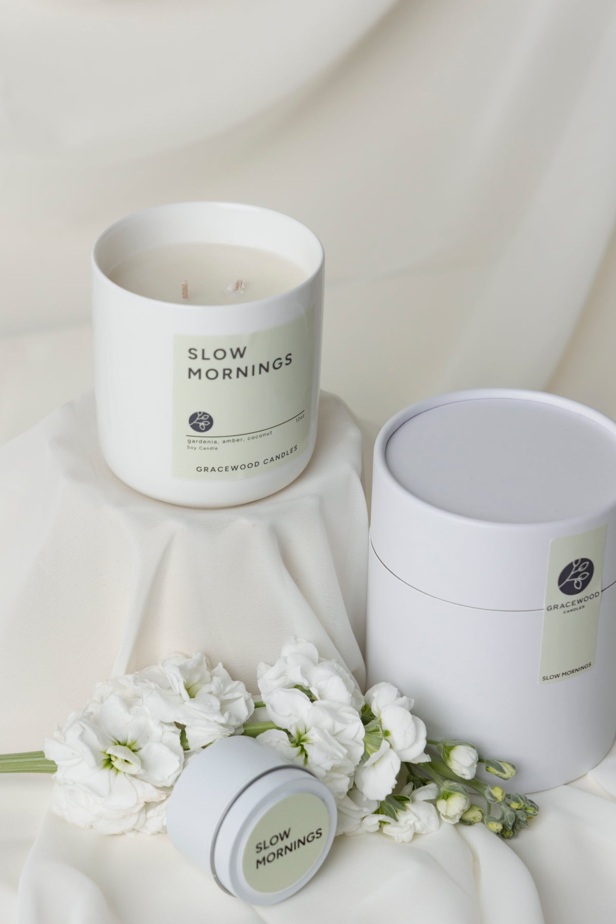 Candle labeled 'Slow Mornings' with a matching container on a soft fabric background.
