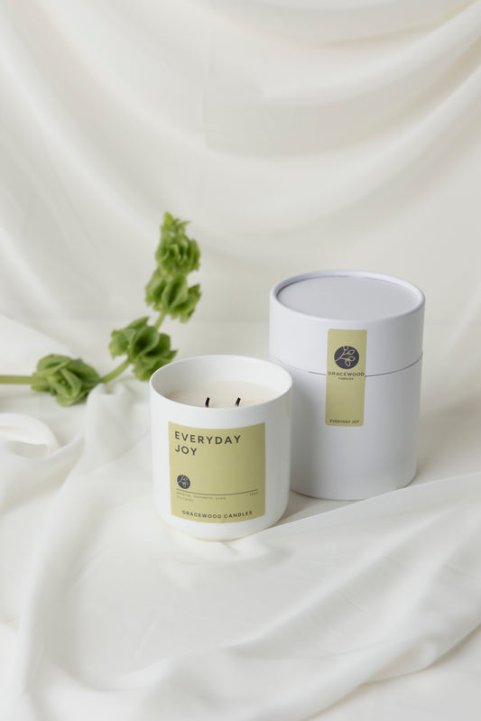 Candle labeled 'Everyday Joy' with a matching white container on a white fabric background